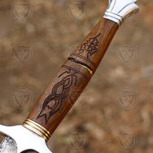 Puede incluir: Primer plano de una empu&ntilde;adura de espada con intrincadas tallas de madera. La empu&ntilde;adura es de madera marr&oacute;n oscuro con una guarda y una hoja de metal plateado. La madera presenta tallas detalladas de un &aacute;rbol y un dise&ntilde;o abstracto.