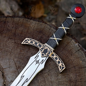 May include: A decorative sword with a silver blade and black handle. The handle is wrapped with white string and gold accents. The pommel features a red gem. The sword rests on a wooden surface.