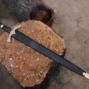 May include: A decorative sword with a black sheath, displayed on a wooden log. The sword has a gold-coloured crossguard and a red gem on the pommel. The handle is wrapped in black leather.