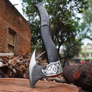 May include: A black axe with a curved handle and a silver blade. The handle is wrapped with a light-colored stitching near the top. The blade has a decorative silver band with an intricate design. The axe is resting on a wooden surface.