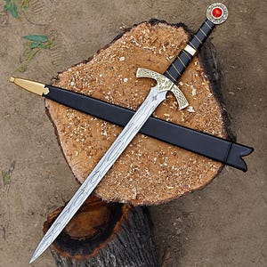 May include: A silver-toned metal sword with a black wrapped handle, a gold-colored crossguard with intricate engravings, and a pommel featuring a red circular stone. The sword is accompanied by a black scabbard with a gold-colored metal tip.