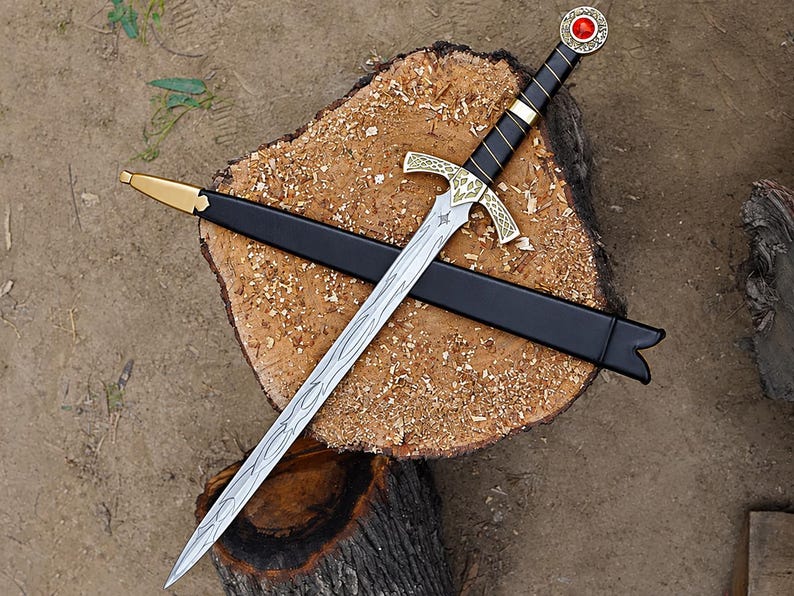 May include: A silver-toned metal sword with a black wrapped handle, a gold-colored crossguard with intricate engravings, and a pommel featuring a red circular stone. The sword is accompanied by a black scabbard with a gold-colored metal tip.