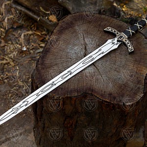 May include: A decorative sword with a silver blade and ornate black and gold hilt, resting on a weathered wooden surface. The blade features a detailed etched design. The hilt is wrapped in black leather with gold accents and a red gem at the pommel.