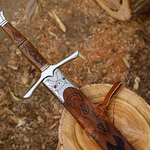 Puede incluir: Una espada de estilo medieval con empu&ntilde;adura de madera y hoja plateada. La empu&ntilde;adura est&aacute; tallada con un patr&oacute;n decorativo. La espada est&aacute; enfundada en una vaina de cuero marr&oacute;n. La espada descansa sobre una superficie de madera.