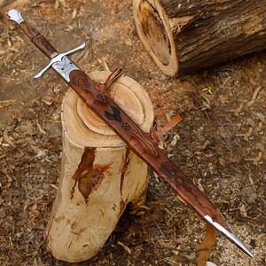 Puede incluir: Una espada de estilo medieval con empu&ntilde;adura de madera y guarda y hoja plateadas. La espada est&aacute; enfundada en una vaina de cuero marr&oacute;n con grabados decorativos. La espada descansa sobre un toc&oacute;n de madera.