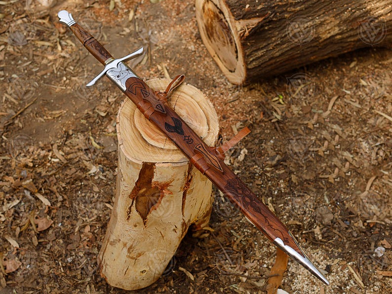 Puede incluir: Una espada de estilo medieval con empu&ntilde;adura de madera y guarda y hoja plateadas. La espada est&aacute; enfundada en una vaina de cuero marr&oacute;n con grabados decorativos. La espada descansa sobre un toc&oacute;n de madera.