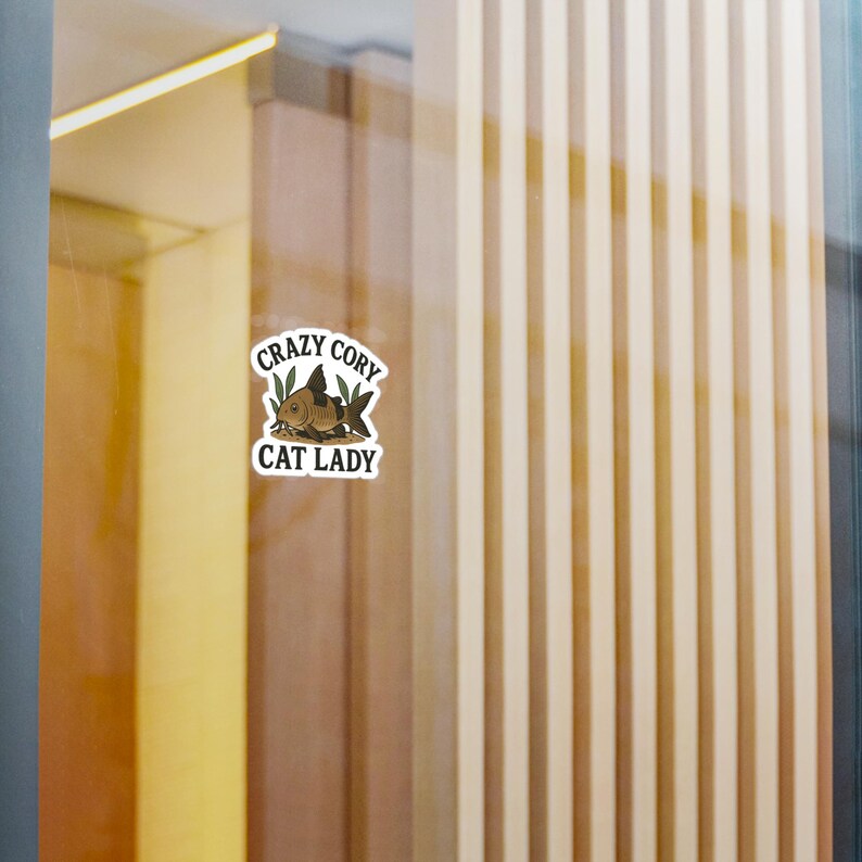 May include: A sticker with the text "Crazy Cory Cat Lady" in a white oval. The sticker features a cartoon illustration of a fish with aquatic plants. The fish is brown with black accents.