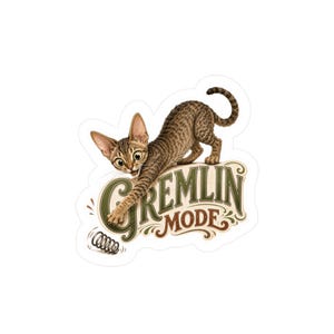 May include: A die-cut sticker featuring a brown tabby cat in a pouncing pose with wide eyes. The cat reaches toward a small metal spring. Below the cat, the text GREMLIN MODE is written in a decorative, vintage-style green and brown font.