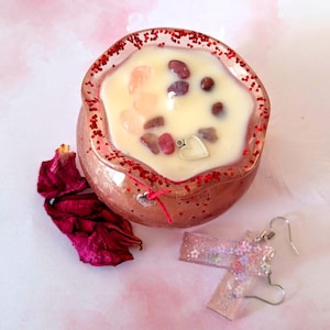 May include: A rose-colored candle with red glitter around the rim, filled with cream-colored wax and gemstones. A small silver heart charm hangs from the candle. A pair of pink, glittery earrings with floral accents are next to the candle. Dried rose petals are scattered nearby.