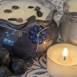 May include: Two decorative candles in blue glass containers with a floral design, filled with white wax and decorative stones. A lit tea light candle and fresh blueberries are in the foreground. The candles have a celestial theme.
