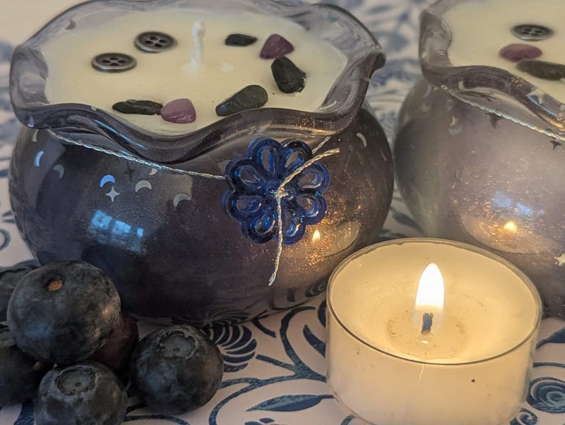 May include: Two decorative candles in blue glass containers with a floral design, filled with white wax and decorative stones. A lit tea light candle and fresh blueberries are in the foreground. The candles have a celestial theme.