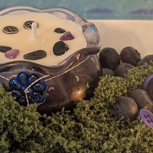May include: A decorative candle in a dark purple glass container with a clear, scalloped rim. The candle is cream-colored and adorned with small stones and buttons. A blue flower and green moss surround the candle, with additional stones and a lit candle in the background.
