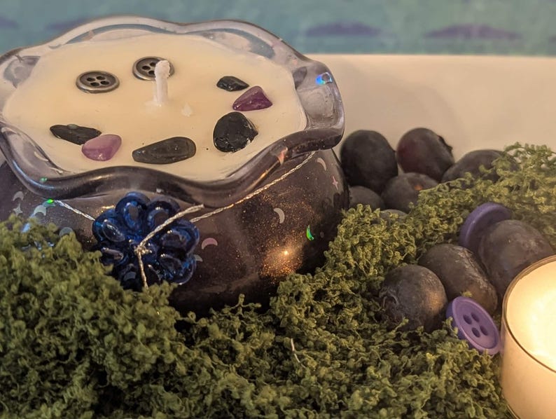 May include: A decorative candle in a dark purple glass container with a clear, scalloped rim. The candle is cream-colored and adorned with small stones and buttons. A blue flower and green moss surround the candle, with additional stones and a lit candle in the background.