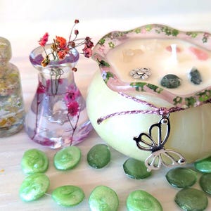 May include: A decorative candle in a round, pale yellow container with a pink floral rim. The candle is adorned with crystals and a silver butterfly charm. Beside it are small glass vases with flowers and decorative green stones.