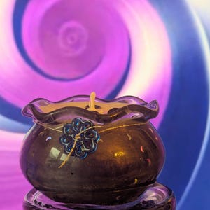 May include: A dark purple candle holder with a wavy rim, holding a lit candle. A blue flower decoration is attached to the front. The background features a swirling pattern of purple and blue.
