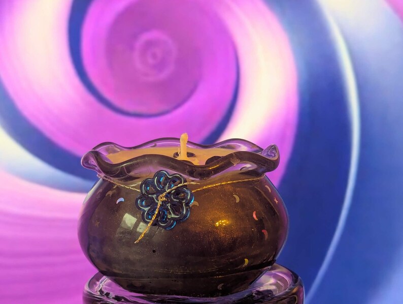 May include: A dark purple candle holder with a wavy rim, holding a lit candle. A blue flower decoration is attached to the front. The background features a swirling pattern of purple and blue.
