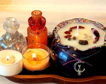 May include: A collection of candles and decorative bottles on a wooden tray. The centerpiece is a black cauldron-shaped candle with a white wax, adorned with crystals and a silver charm. Two small candles and two glitter-filled bottles complete the arrangement.