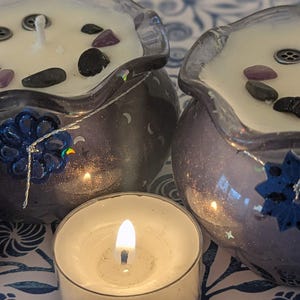 May include: Two decorative candles in purple glass bowls, each with a white wax surface adorned with black and purple stones. A small lit tea light candle is in front. The bowls have blue floral accents and silver string.