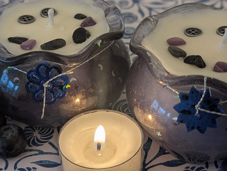 May include: Two decorative candles in purple glass bowls, each with a white wax surface adorned with black and purple stones. A small lit tea light candle is in front. The bowls have blue floral accents and silver string.