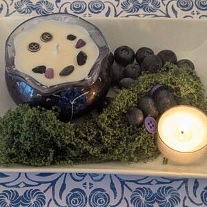 May include: A decorative arrangement on a white tray featuring a dark blue candle with decorative stones and buttons, blueberries, green moss, and a lit tea light candle. The tray sits on a blue and white patterned surface.