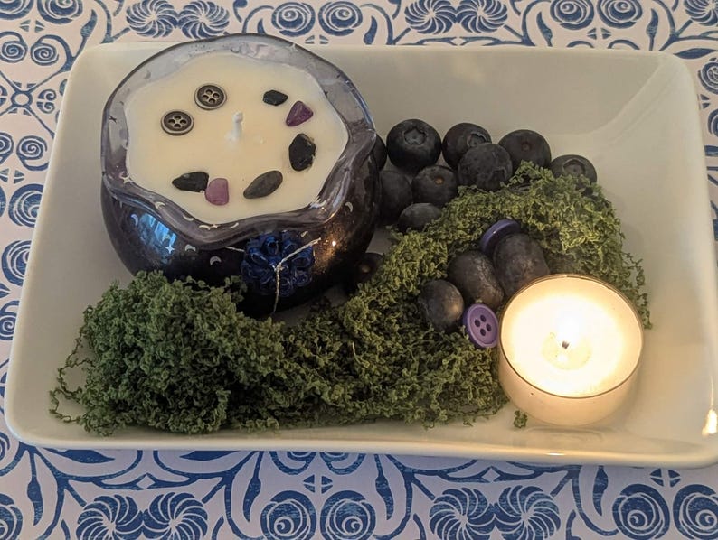 May include: A decorative arrangement on a white tray featuring a dark blue candle with decorative stones and buttons, blueberries, green moss, and a lit tea light candle. The tray sits on a blue and white patterned surface.