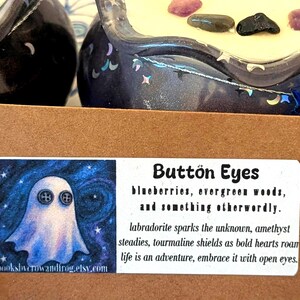 May include: A close-up of two decorative candle holders with a dark blue, starry design. The candles are topped with small stones. A label reads "Button Eyes" with text about the product and a ghost illustration.