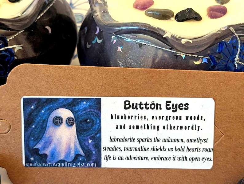 May include: A close-up of two decorative candle holders with a dark blue, starry design. The candles are topped with small stones. A label reads "Button Eyes" with text about the product and a ghost illustration.