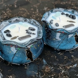 May include: Two blue glass candles with a marbled effect, each with a white wax top. The candles are decorated with black stones, small blue crystals, and a metal charm that reads "Leaves". A black raven charm hangs from the front of each candle.