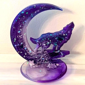 May include: A purple resin sculpture of a howling wolf silhouetted against a crescent moon. The moon and base are filled with glitter and star-shaped accents. The sculpture is approximately 4 inches tall.