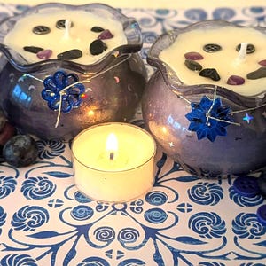 May include: Two purple glass candles with white wax, decorated with stones and buttons, sit on a blue and white patterned surface. A lit tea light and fresh blueberries are in the foreground. Decorative blue flower accents adorn the candle holders.