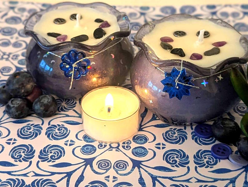 May include: Two purple glass candles with white wax, decorated with stones and buttons, sit on a blue and white patterned surface. A lit tea light and fresh blueberries are in the foreground. Decorative blue flower accents adorn the candle holders.