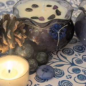 May include: Two decorative candles in dark blue glass containers with a floral design, filled with cream-colored wax and decorative stones. A lit tea light candle, pine cone, and blue buttons are also present, all set on a patterned surface.