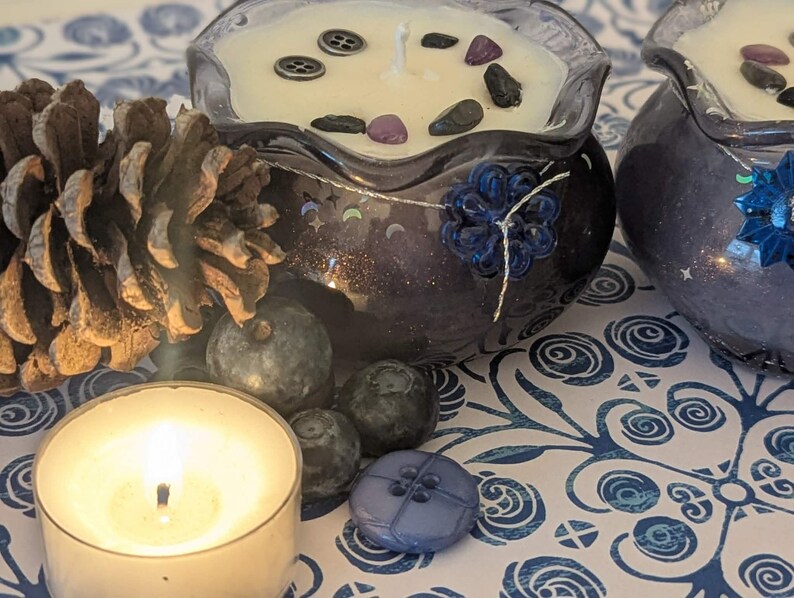 May include: Two decorative candles in dark blue glass containers with a floral design, filled with cream-colored wax and decorative stones. A lit tea light candle, pine cone, and blue buttons are also present, all set on a patterned surface.
