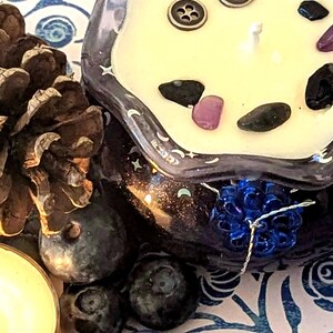 May include: A decorative candle in a dark glass container with a white wax top, adorned with small gemstones and a button. The candle sits next to a pine cone, blueberries, and a lit tea light on a patterned surface.