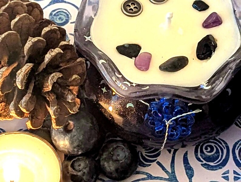 May include: A decorative candle in a dark glass container with a white wax top, adorned with small gemstones and a button. The candle sits next to a pine cone, blueberries, and a lit tea light on a patterned surface.