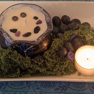 May include: A candle in a dark blue glass container with decorative stones and buttons, surrounded by green moss, blueberries, and a lit votive candle. The arrangement is on a white rectangular dish.