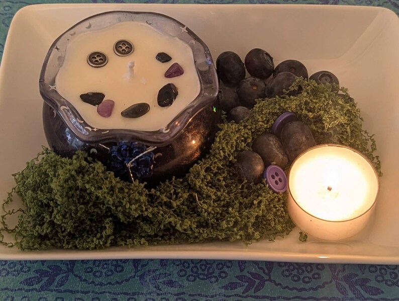 May include: A candle in a dark blue glass container with decorative stones and buttons, surrounded by green moss, blueberries, and a lit votive candle. The arrangement is on a white rectangular dish.