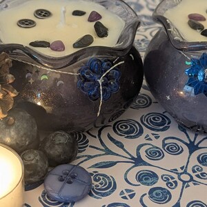 May include: Two decorative candles in dark blue glass bowls, filled with cream-colored wax and adorned with decorative stones and buttons. A pine cone, blueberries, and a lit candle are arranged nearby, creating a cozy, nature-inspired theme.