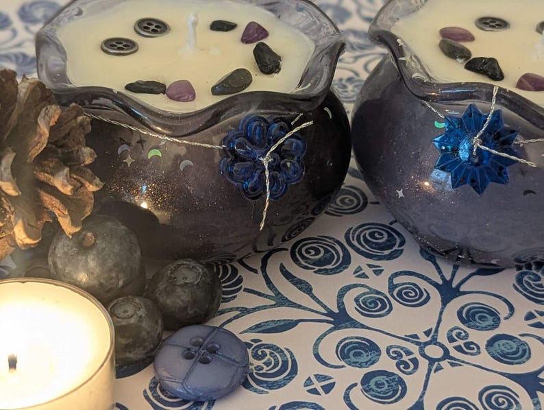 May include: Two decorative candles in dark blue glass bowls, filled with cream-colored wax and adorned with decorative stones and buttons. A pine cone, blueberries, and a lit candle are arranged nearby, creating a cozy, nature-inspired theme.