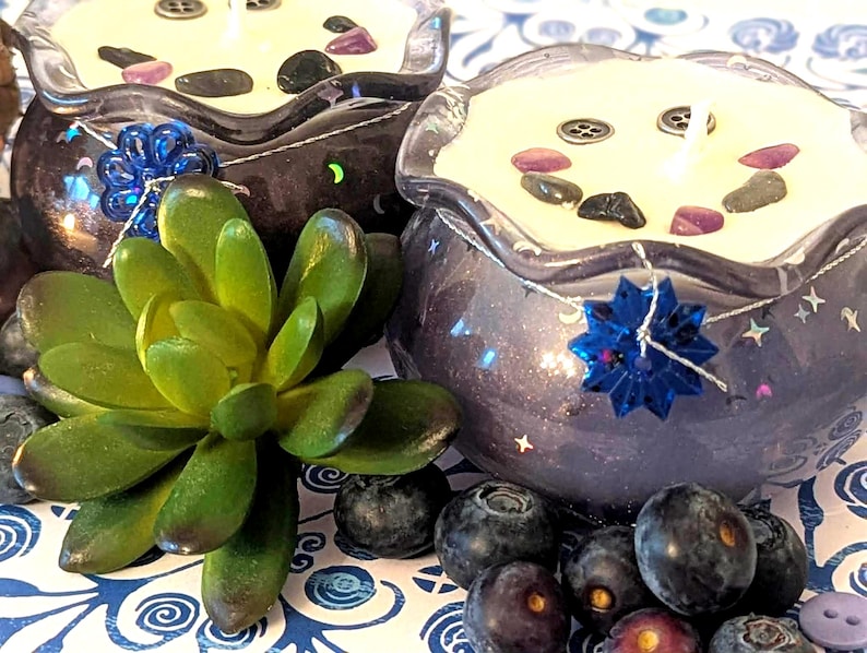 May include: Two decorative candles in dark blue glass bowls with a starry design. Each candle has a white wax top with decorative stones and a wick. A blue flower and a succulent plant are in the foreground, with blueberries scattered around.
