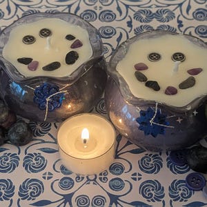 May include: Two decorative candles in purple glass containers with white wax, adorned with stones and buttons. A lit tea light candle sits in front of the candles. Blueberries and buttons are scattered around the candles on a blue and white patterned surface.
