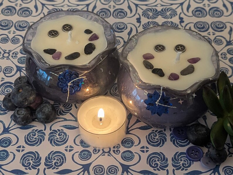 May include: Two decorative candles in purple glass containers with white wax, adorned with stones and buttons. A lit tea light candle sits in front of the candles. Blueberries and buttons are scattered around the candles on a blue and white patterned surface.