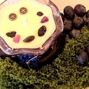 May include: A decorative candle in a dark glass container, surrounded by green moss, blueberries, and purple buttons. The candle has a light yellow wax with decorative stones and buttons.
