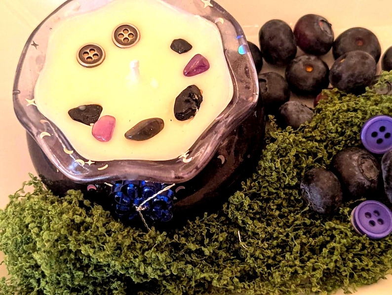 May include: A decorative candle in a dark glass container, surrounded by green moss, blueberries, and purple buttons. The candle has a light yellow wax with decorative stones and buttons.