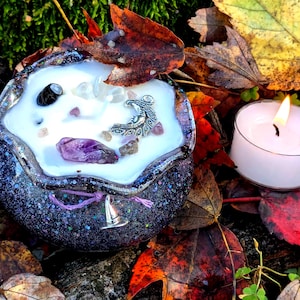 May include: A round, dark purple glittery candle holder with a white candle, decorated with crystals and a silver charm. A lit white tea light candle sits nearby. The scene is set among colorful autumn leaves.