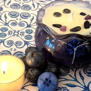 May include: A lit tealight candle and a blueberry-scented candle in a dark blue decorative bowl. The candle is decorated with small stones and buttons. Fresh blueberries and a blue button are also present on a blue and white patterned surface.