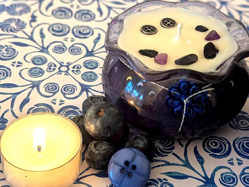 May include: A lit tealight candle and a blueberry-scented candle in a dark blue decorative bowl. The candle is decorated with small stones and buttons. Fresh blueberries and a blue button are also present on a blue and white patterned surface.