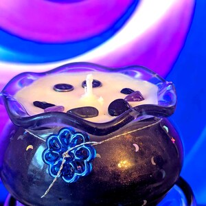 May include: A dark blue candle in a glass container with a ruffled rim. The candle is decorated with small stones and a blue flower. The background is a swirl of blue, purple, and white.