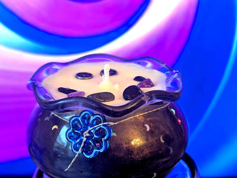 May include: A dark blue candle in a glass container with a ruffled rim. The candle is decorated with small stones and a blue flower. The background is a swirl of blue, purple, and white.