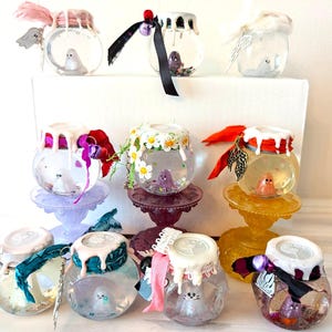 May include: A collection of decorative glass jars with various embellishments. Each jar contains a small figurine and is topped with a white, dripped-wax effect. The jars are adorned with ribbons, feathers, and floral accents, and sit on decorative stands.